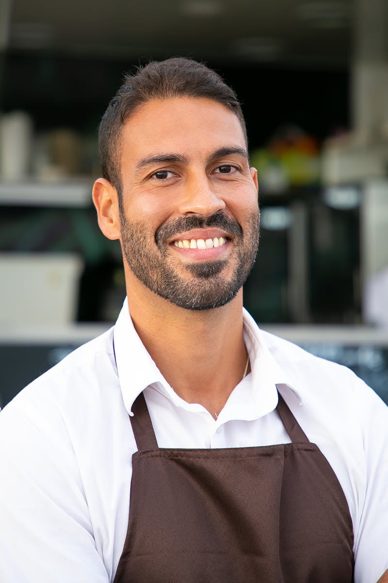 Smiling male café worker in apron, standing confidently outdoors in a business setting.